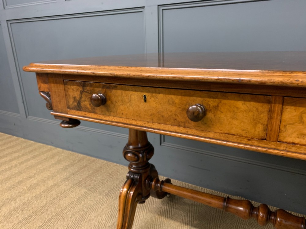 A Victorian burr walnut library table featuring a polished rectangular top with rounded corners, frieze drawers with wooden turned knobs, and twin carved end supports joined by a turned stretcher. View VII