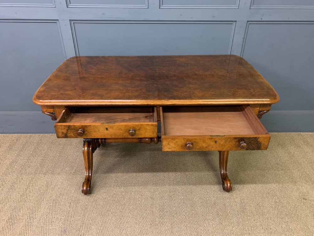 A Victorian burr walnut library table featuring a polished rectangular top with rounded corners, frieze drawers with wooden turned knobs, and twin carved end supports joined by a turned stretcher. View IX