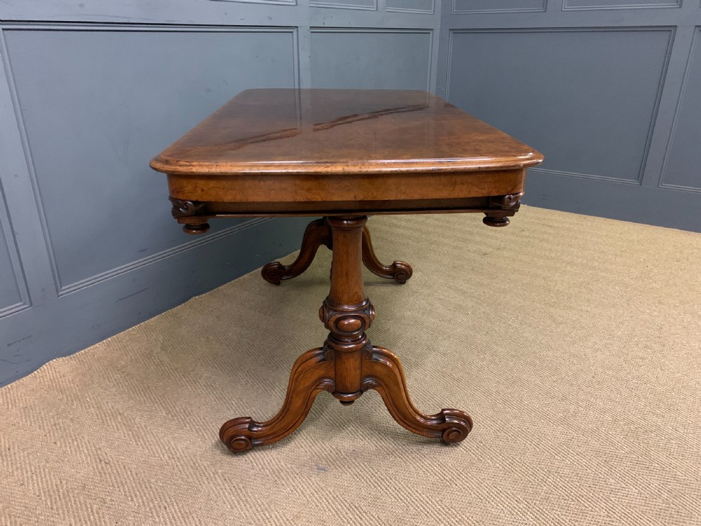 A Victorian burr walnut library table featuring a polished rectangular top with rounded corners, frieze drawers with wooden turned knobs, and twin carved end supports joined by a turned stretcher. View X