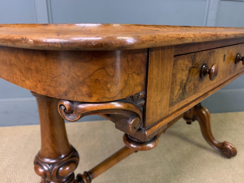 A Victorian burr walnut library table featuring a polished rectangular top with rounded corners, frieze drawers with wooden turned knobs, and twin carved end supports joined by a turned stretcher. View XVI