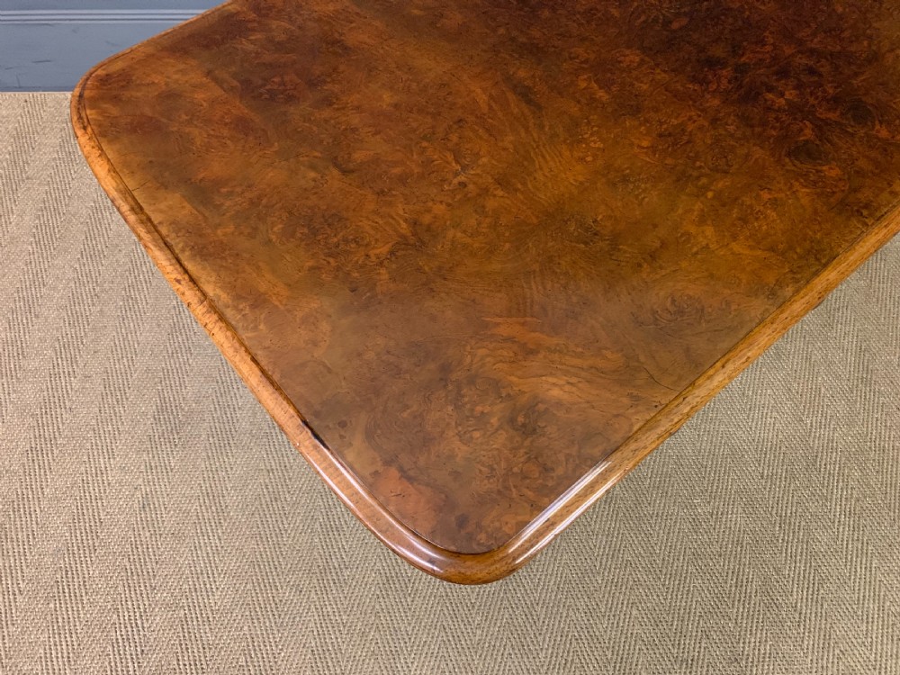 A Victorian burr walnut library table featuring a polished rectangular top with rounded corners, frieze drawers with wooden turned knobs, and twin carved end supports joined by a turned stretcher. View XVII