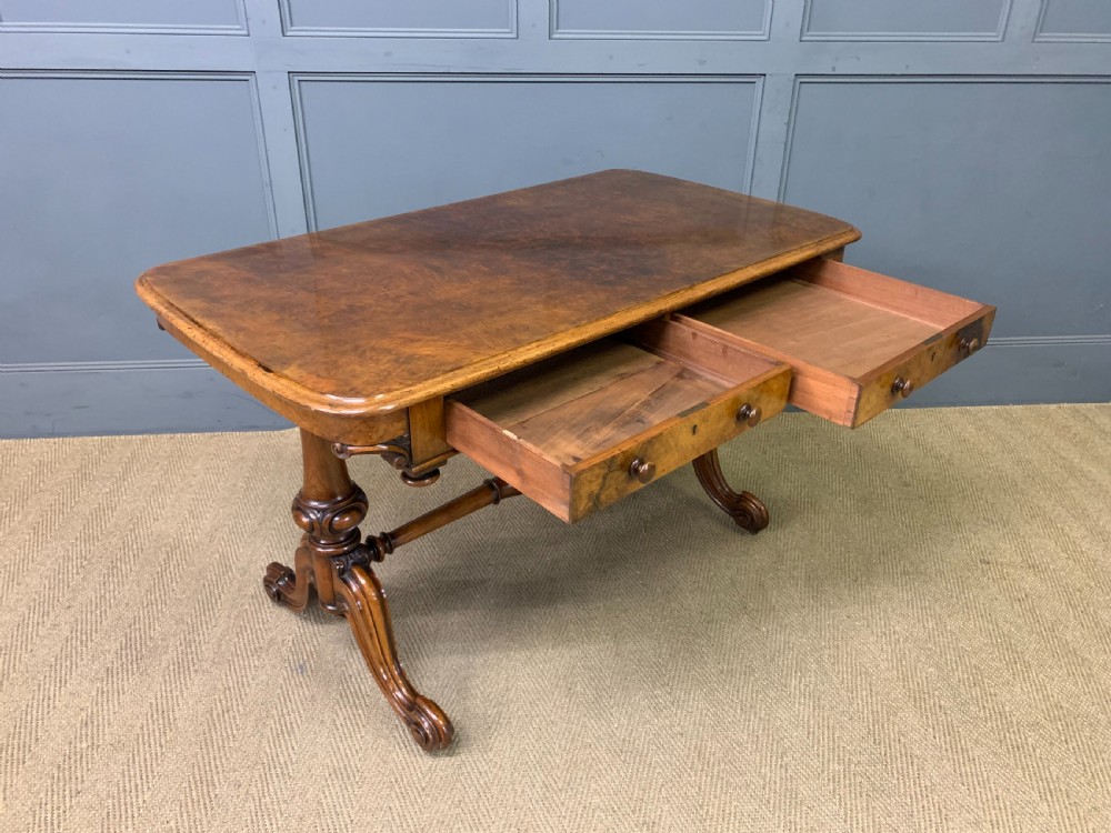 A Victorian burr walnut library table featuring a polished rectangular top with rounded corners, frieze drawers with wooden turned knobs, and twin carved end supports joined by a turned stretcher. View XVIII