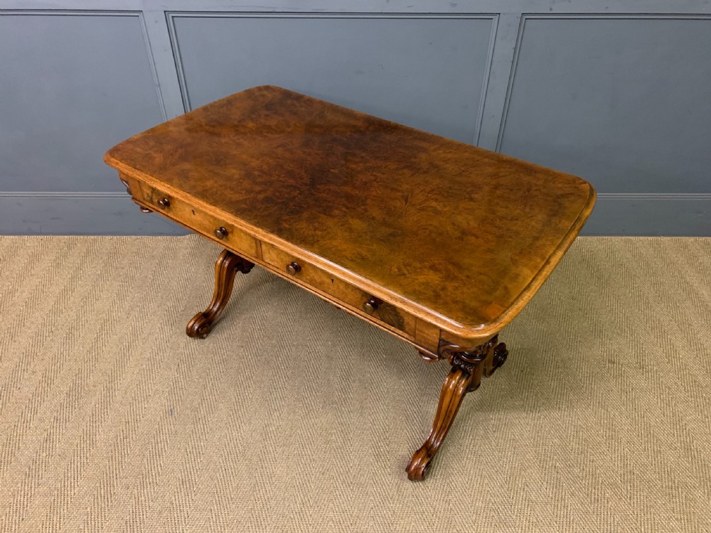 A Victorian burr walnut library table featuring a polished rectangular top with rounded corners, frieze drawers with wooden turned knobs, and twin carved end supports joined by a turned stretcher. View XIX