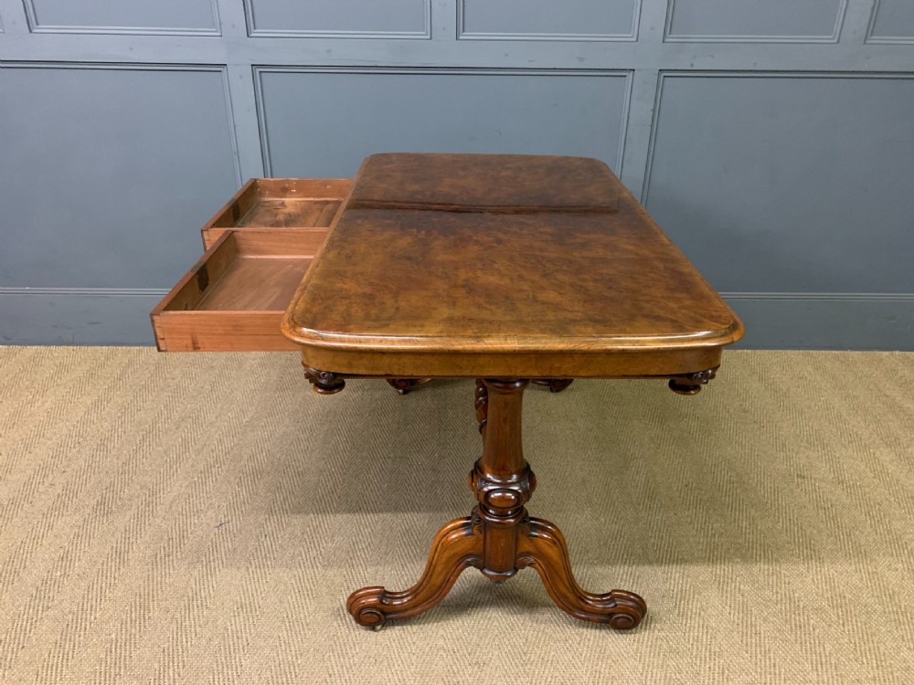 A Victorian burr walnut library table featuring a polished rectangular top with rounded corners, frieze drawers with wooden turned knobs, and twin carved end supports joined by a turned stretcher. View XX