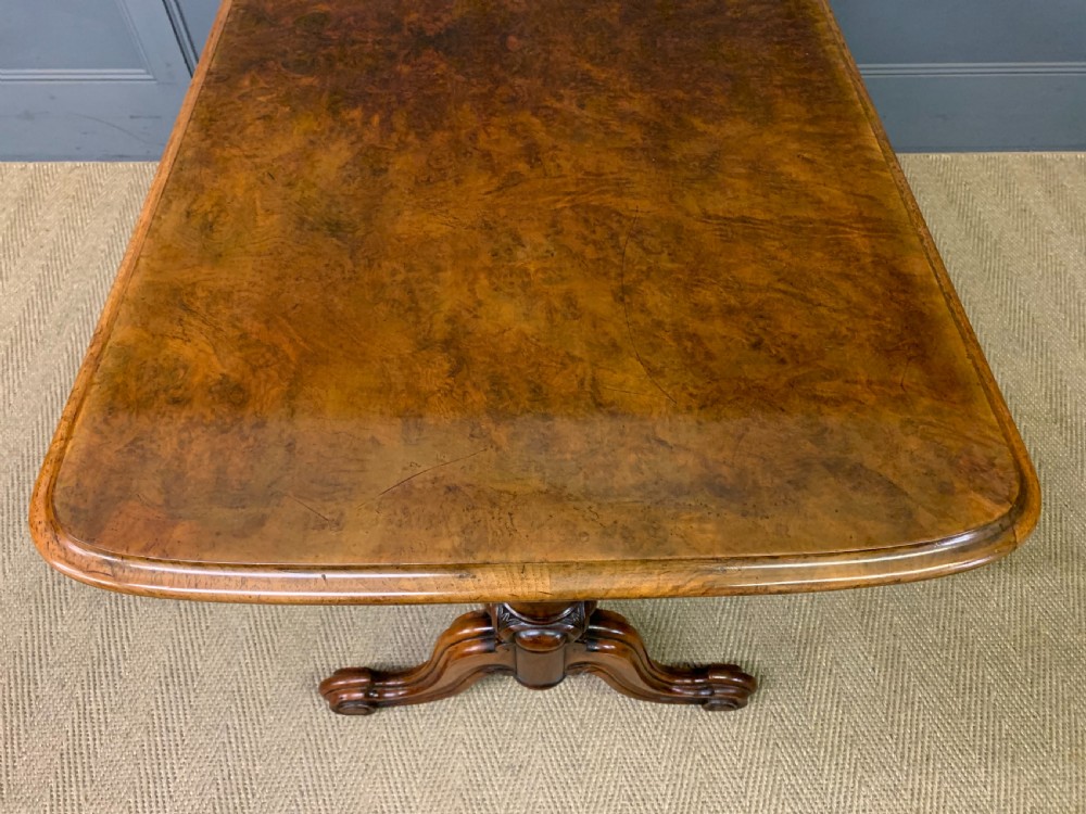 A Victorian burr walnut library table featuring a polished rectangular top with rounded corners, frieze drawers with wooden turned knobs, and twin carved end supports joined by a turned stretcher. View XXI
