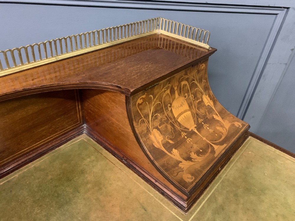 An Edwardian mahogany writing desk featuring a green leather surface, brass gallery rail, marquetry sloped panels, and drawer pedestals on tapered legs. View XVII