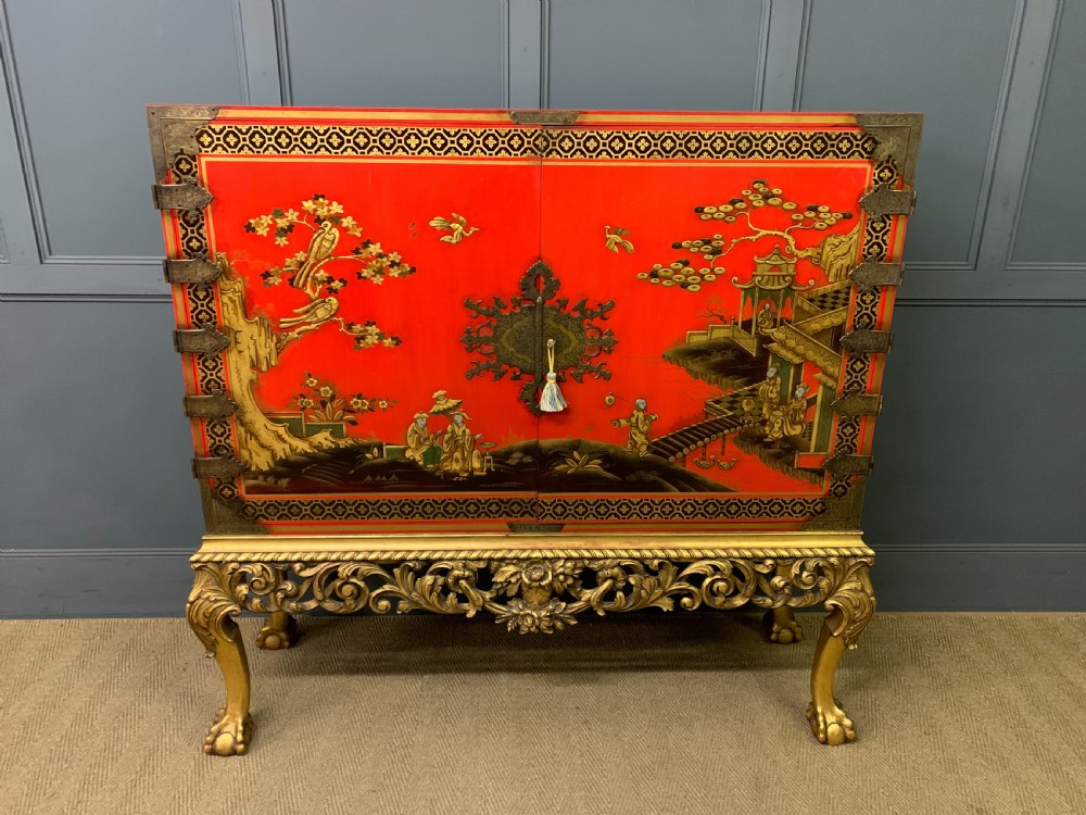 A bright red lacquered Chinoiserie cabinet with gold hand-painted figures and landscapes, mounted on an ornate carved giltwood stand with cabriole legs.