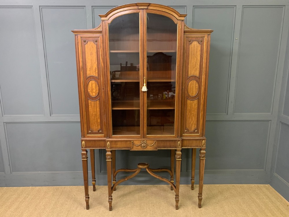 An antique mahogany display cabinet featuring glazed centre doors, side inlaid panels, fluted legs, and an arched top.