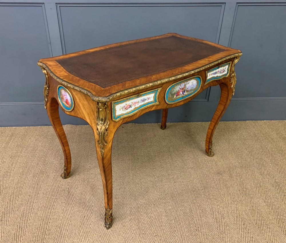 An antique Louis XV style French tulipwood bureau plat featuring a leather-inset top, gilt-bronze mounts, cabriole legs, and hand-painted porcelain plaques.