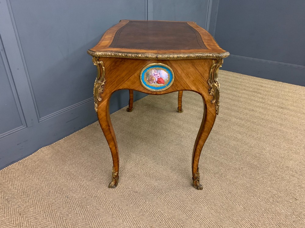 An antique Louis XV style French tulipwood bureau plat featuring a leather-inset top, gilt-bronze mounts, cabriole legs, and hand-painted porcelain plaques. View X