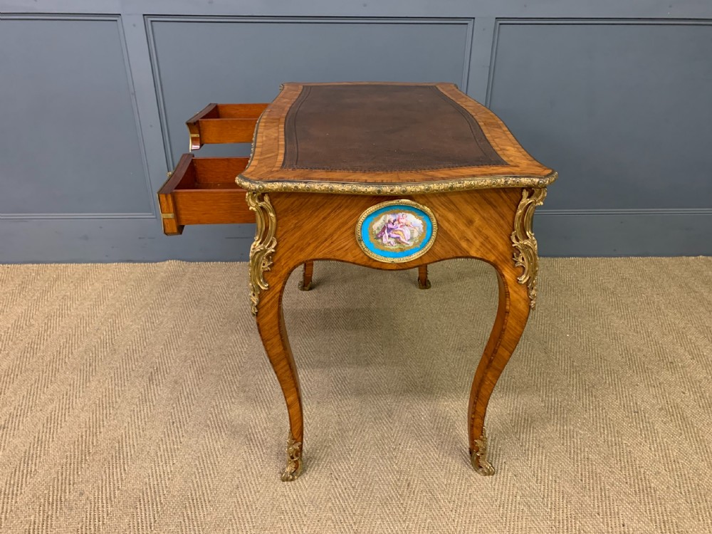 An antique Louis XV style French tulipwood bureau plat featuring a leather-inset top, gilt-bronze mounts, cabriole legs, and hand-painted porcelain plaques. View XIX