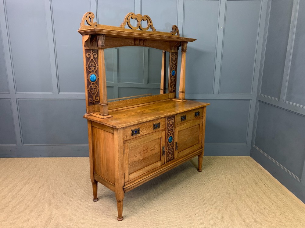 An Arts and Crafts golden oak sideboard featuring a mirrored superstructure with carved fretwork and blue ceramic cabochons above two drawers and two cupboard doors.