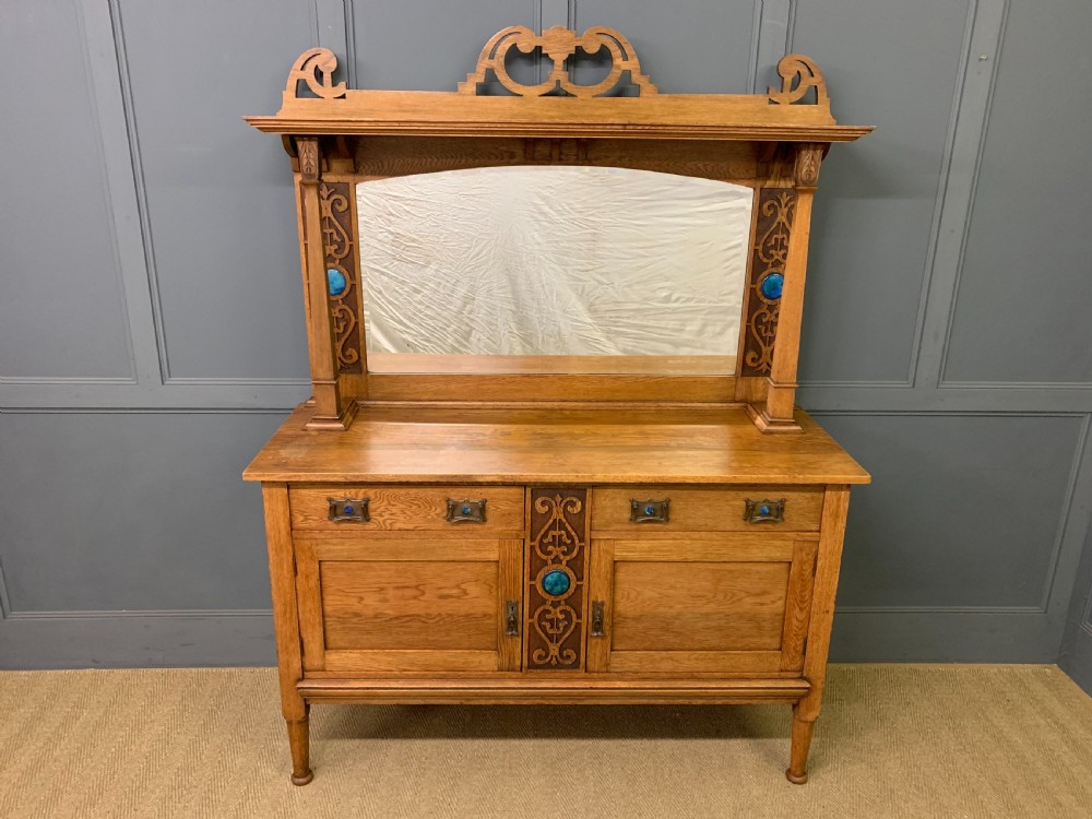 An Arts and Crafts golden oak sideboard featuring a mirrored superstructure with carved fretwork and blue ceramic cabochons above two drawers and two cupboard doors. View II