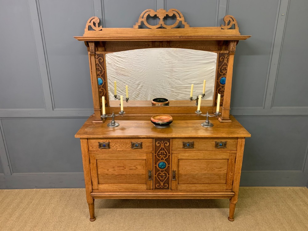 An Arts and Crafts golden oak sideboard featuring a mirrored superstructure with carved fretwork and blue ceramic cabochons above two drawers and two cupboard doors. View III
