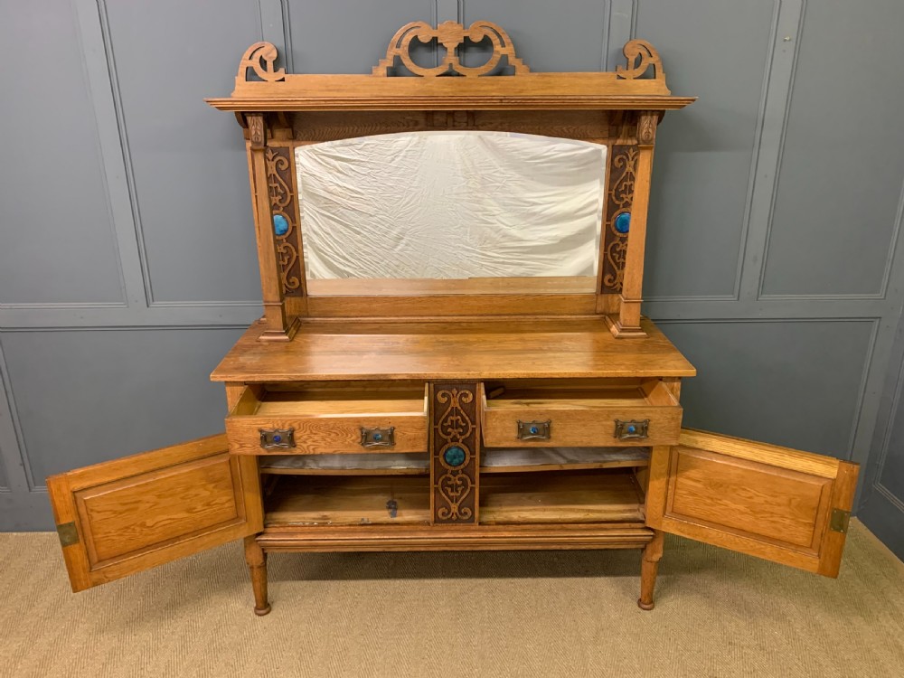 An Arts and Crafts golden oak sideboard featuring a mirrored superstructure with carved fretwork and blue ceramic cabochons above two drawers and two cupboard doors. View IV
