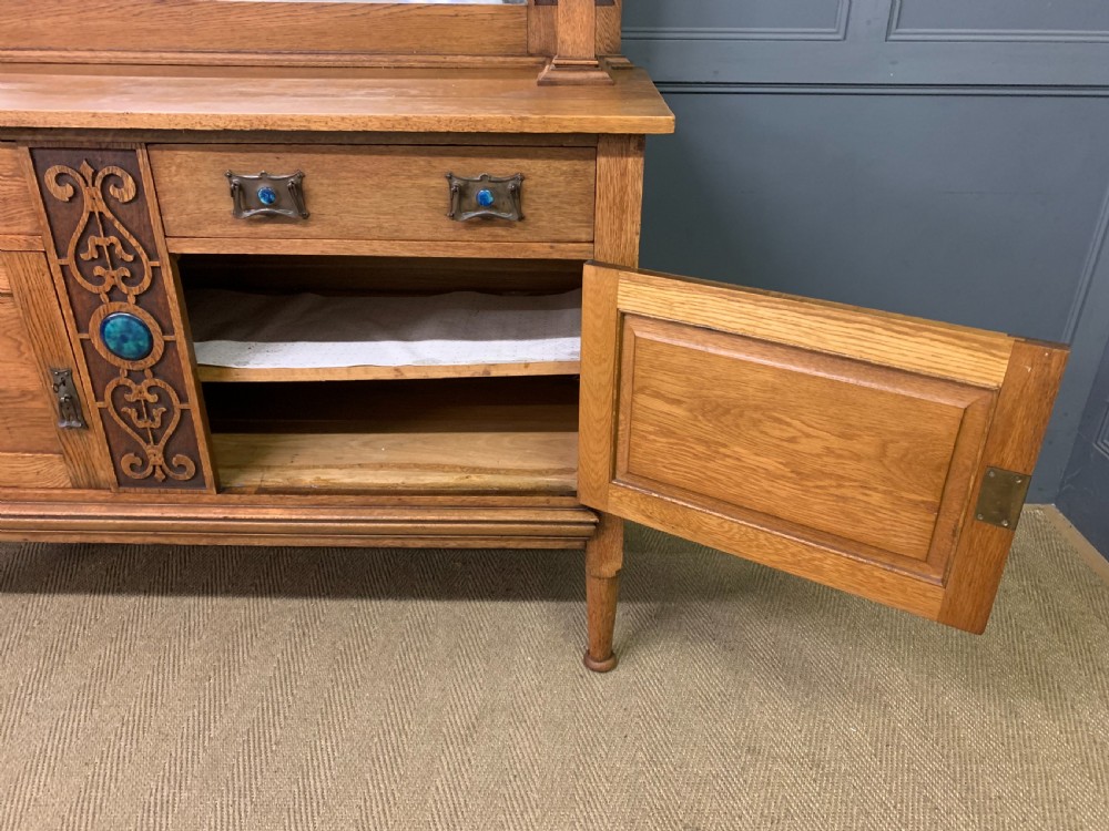 An Arts and Crafts golden oak sideboard featuring a mirrored superstructure with carved fretwork and blue ceramic cabochons above two drawers and two cupboard doors. View V