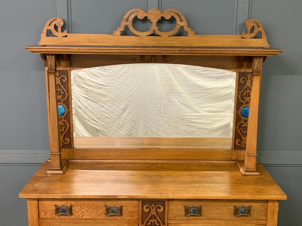 An Arts and Crafts golden oak sideboard featuring a mirrored superstructure with carved fretwork and blue ceramic cabochons above two drawers and two cupboard doors. View VI