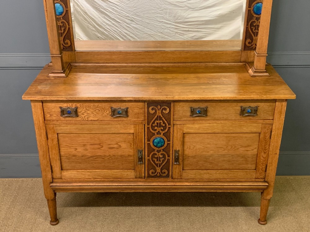 An Arts and Crafts golden oak sideboard featuring a mirrored superstructure with carved fretwork and blue ceramic cabochons above two drawers and two cupboard doors. View VII