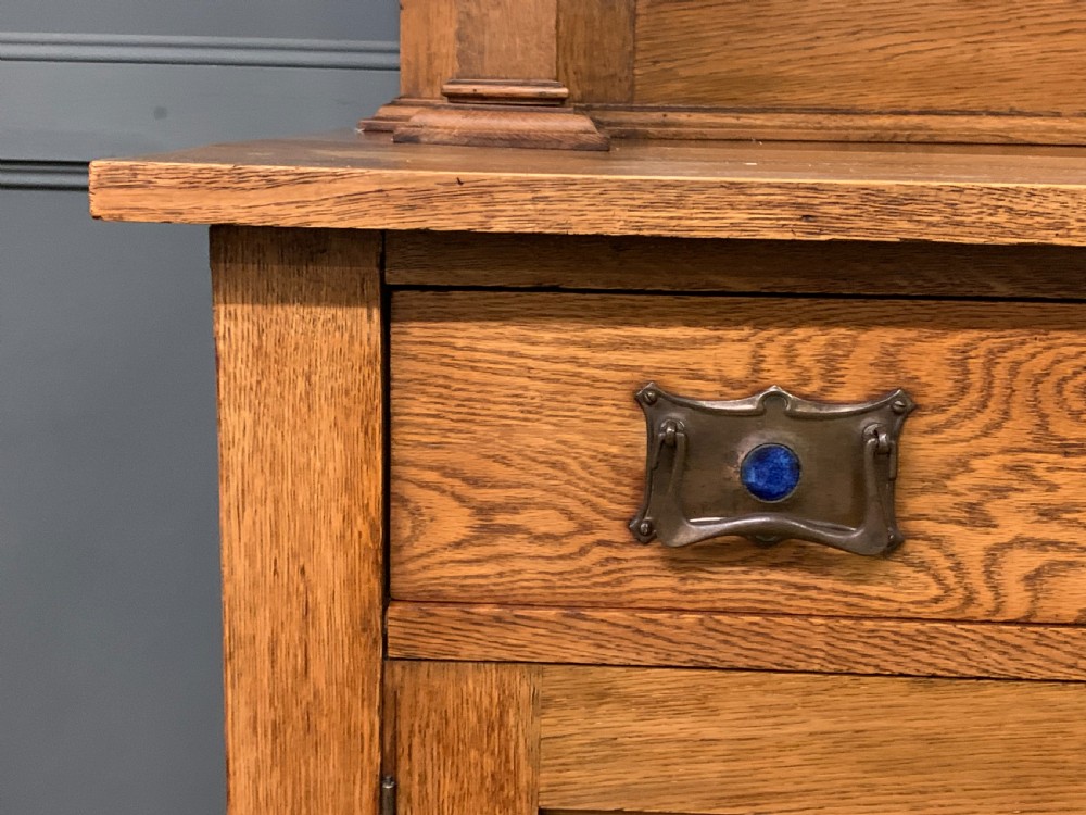 An Arts and Crafts golden oak sideboard featuring a mirrored superstructure with carved fretwork and blue ceramic cabochons above two drawers and two cupboard doors. View XV