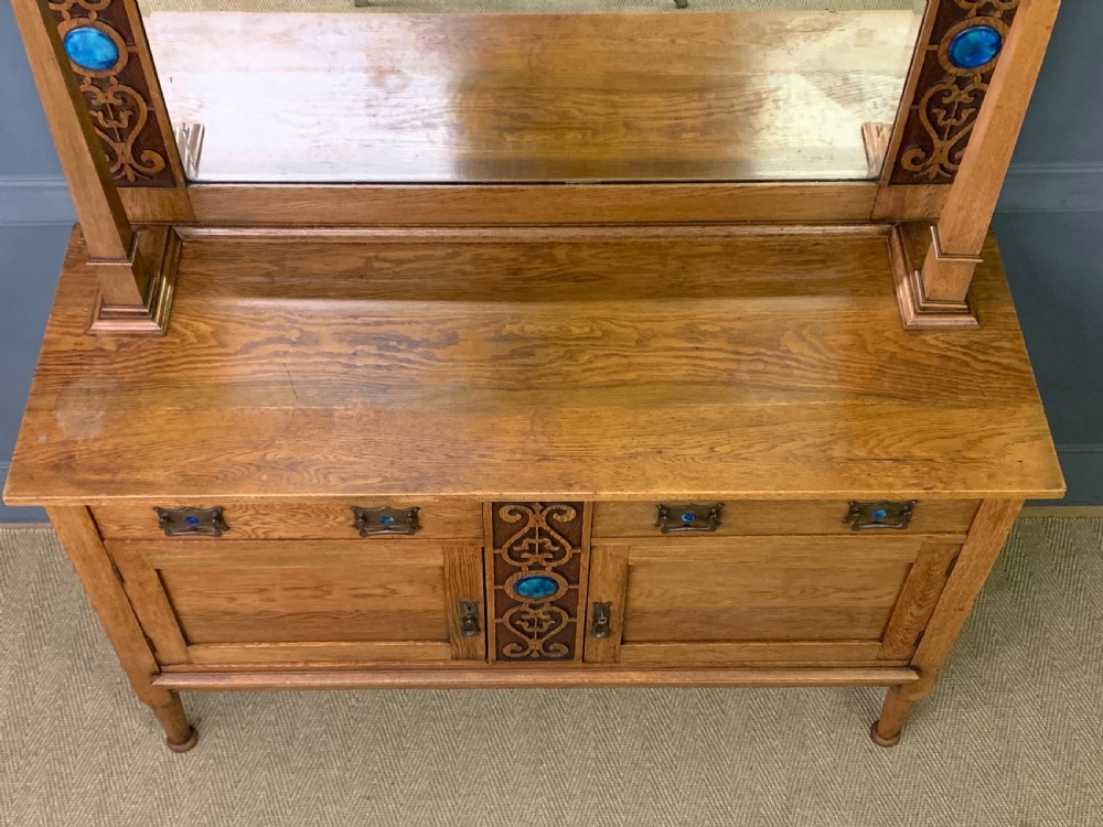 An Arts and Crafts golden oak sideboard featuring a mirrored superstructure with carved fretwork and blue ceramic cabochons above two drawers and two cupboard doors. View XXII