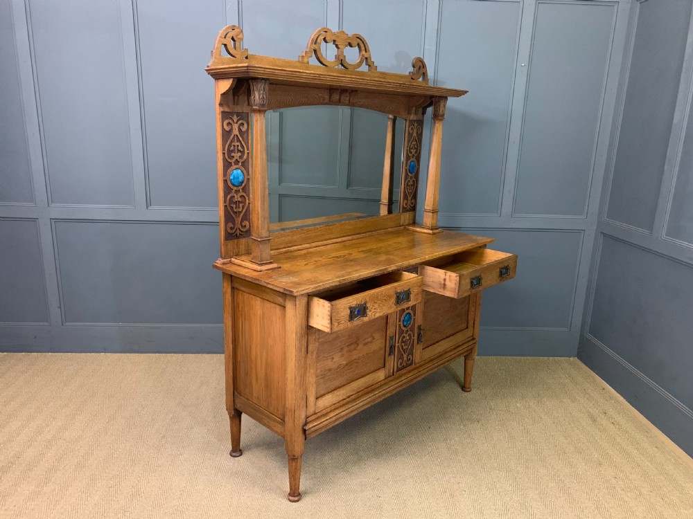 An Arts and Crafts golden oak sideboard featuring a mirrored superstructure with carved fretwork and blue ceramic cabochons above two drawers and two cupboard doors. View XXVI