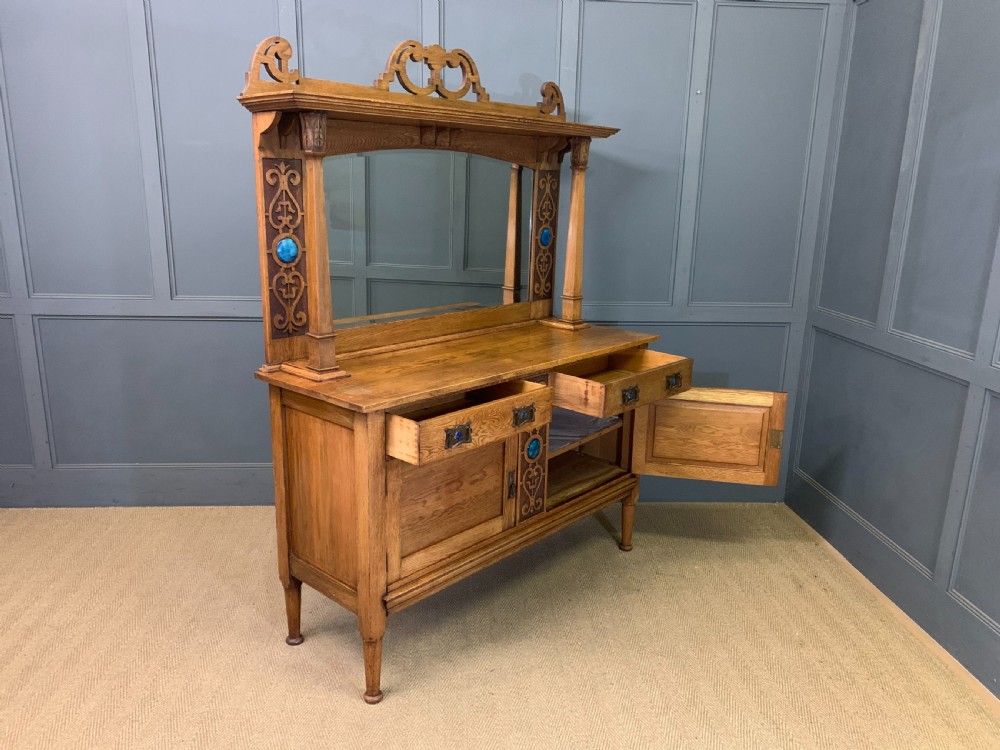 An Arts and Crafts golden oak sideboard featuring a mirrored superstructure with carved fretwork and blue ceramic cabochons above two drawers and two cupboard doors. View XXVII