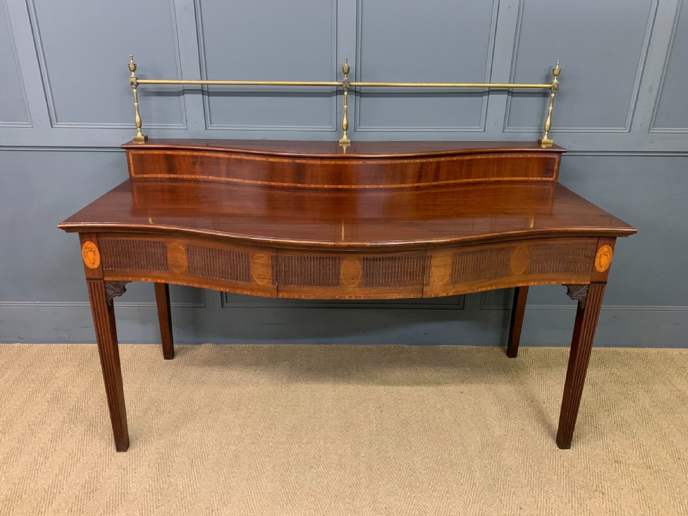 A George III mahogany serving table featuring a serpentine front, inlaid fluted detailing, square tapered legs, and a brass gallery rail.