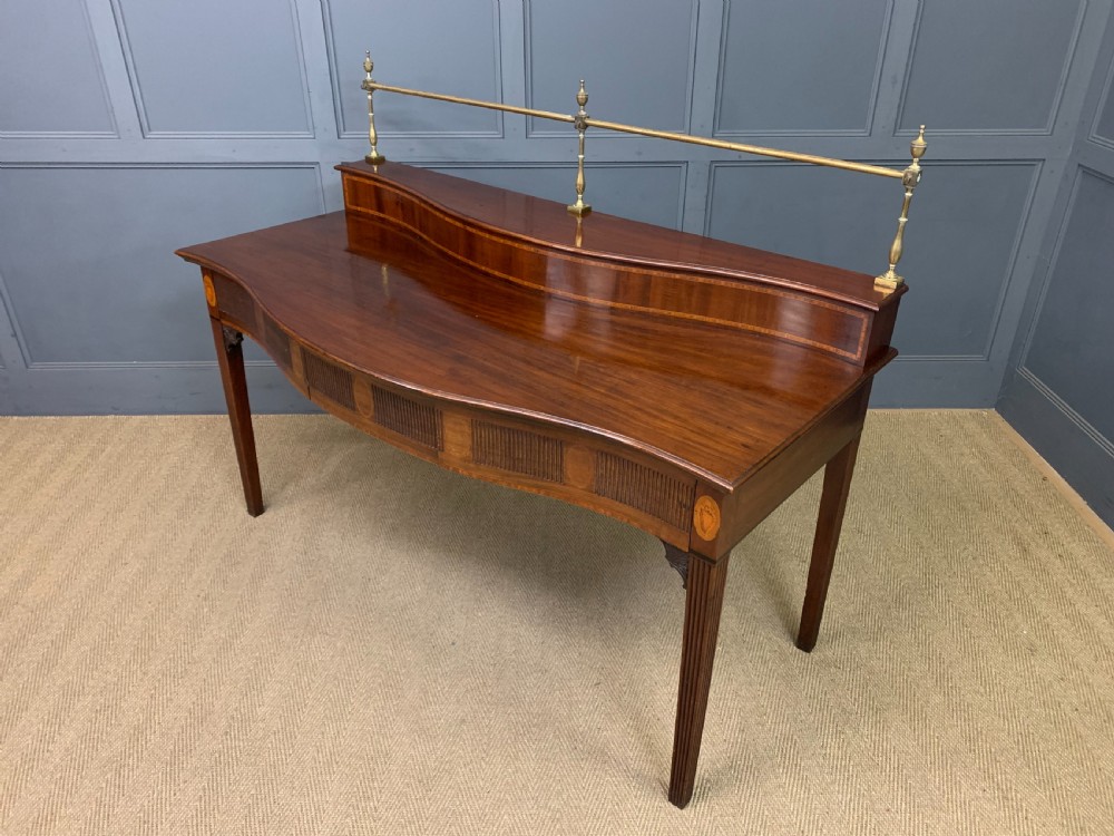 A George III mahogany serving table featuring a serpentine front, inlaid fluted detailing, square tapered legs, and a brass gallery rail. View XIX