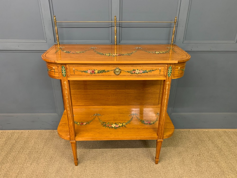 A painted satinwood side table featuring hand-painted floral swags, a single drawer, a lower shelf, tapered square legs, and a raised brass gallery rail.