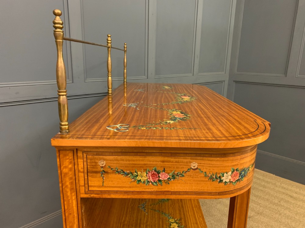 A painted satinwood side table featuring hand-painted floral swags, a single drawer, a lower shelf, tapered square legs, and a raised brass gallery rail. View XVII