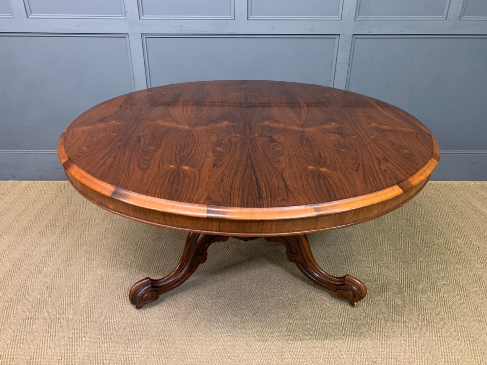 An oval Victorian rosewood loo table featuring a book-matched veneered top, crossbanded edge, and a carved central pedestal with four splayed legs on castors.