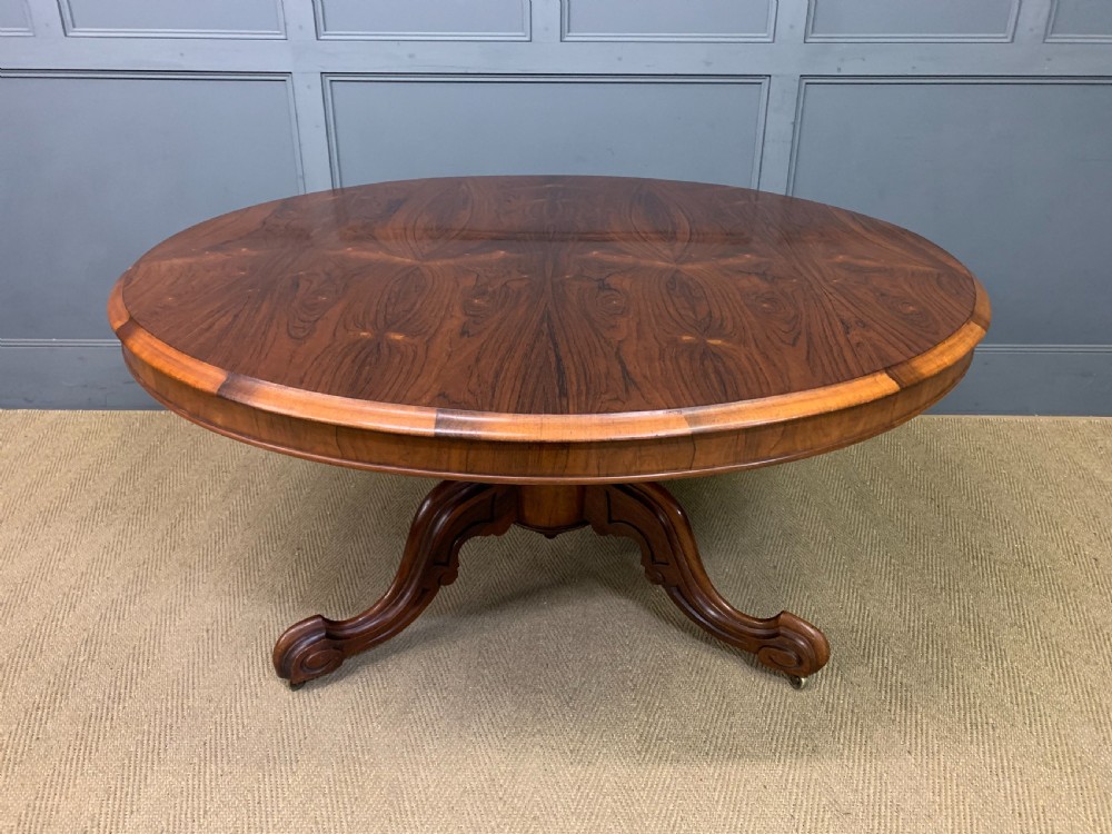 An oval Victorian rosewood loo table featuring a book-matched veneered top, crossbanded edge, and a carved central pedestal with four splayed legs on castors. View II