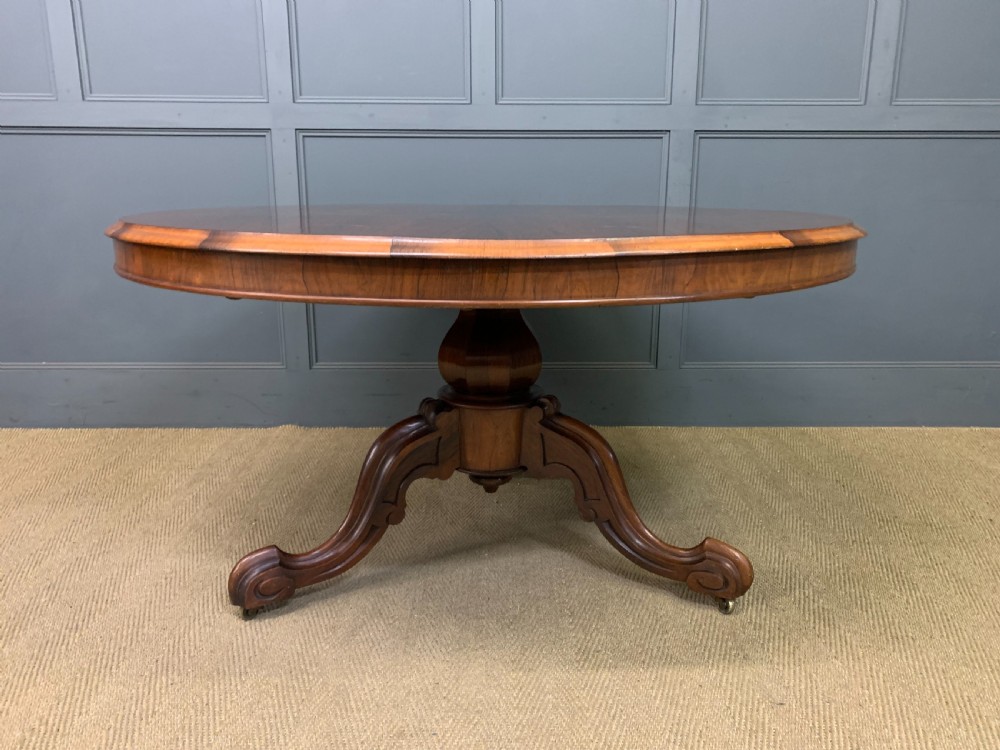 An oval Victorian rosewood loo table featuring a book-matched veneered top, crossbanded edge, and a carved central pedestal with four splayed legs on castors. View III