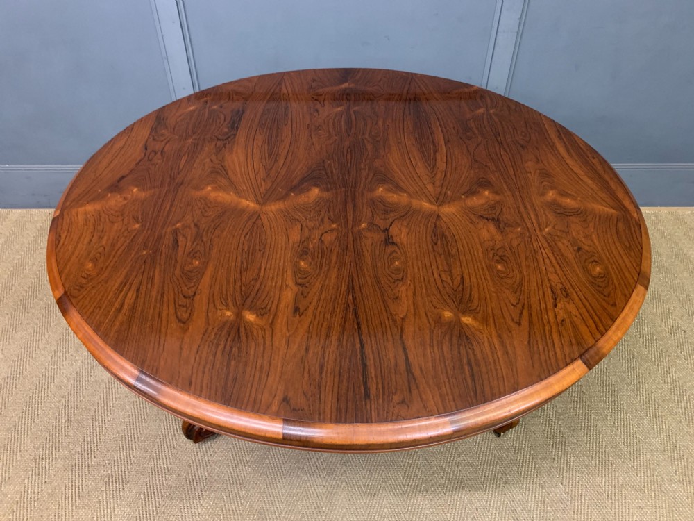 An oval Victorian rosewood loo table featuring a book-matched veneered top, crossbanded edge, and a carved central pedestal with four splayed legs on castors. View IV