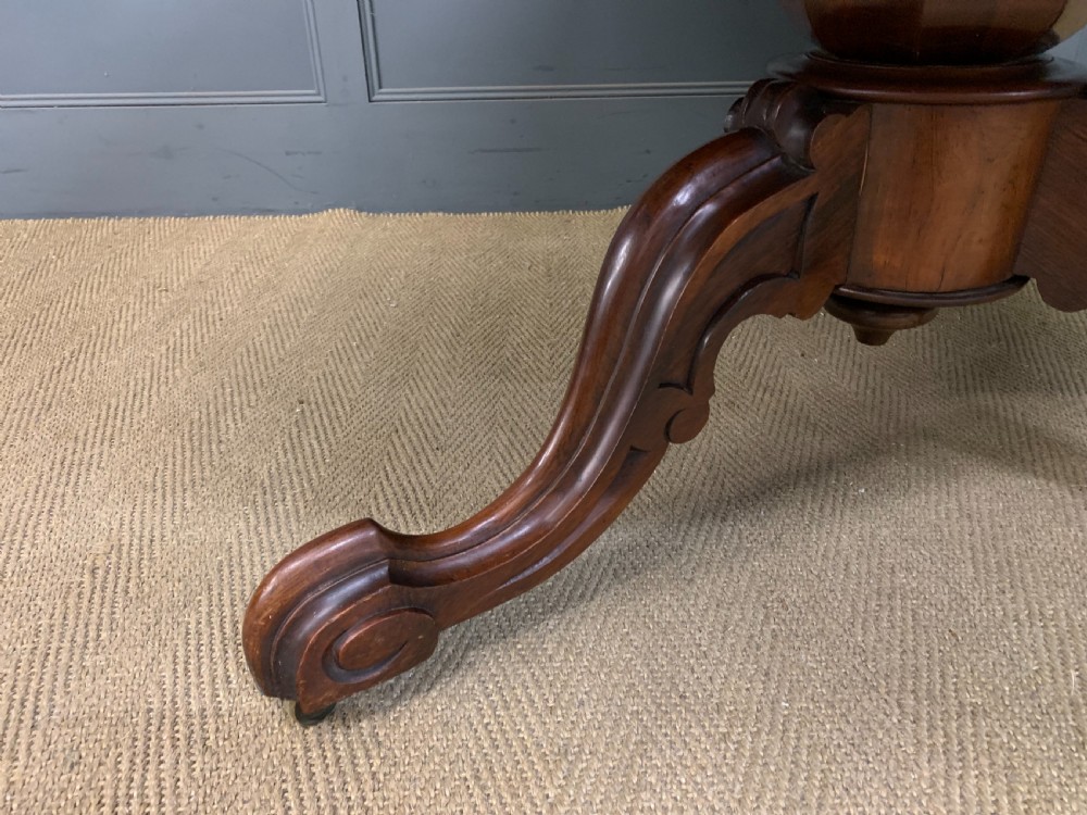 An oval Victorian rosewood loo table featuring a book-matched veneered top, crossbanded edge, and a carved central pedestal with four splayed legs on castors. View V