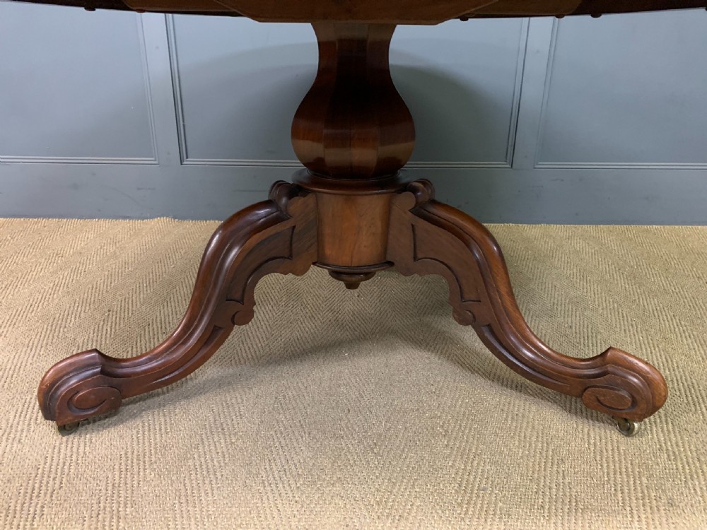 An oval Victorian rosewood loo table featuring a book-matched veneered top, crossbanded edge, and a carved central pedestal with four splayed legs on castors. View VI