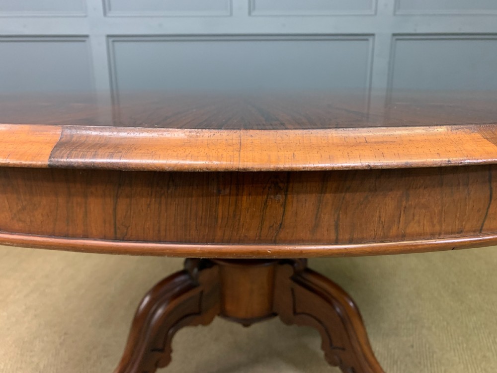 An oval Victorian rosewood loo table featuring a book-matched veneered top, crossbanded edge, and a carved central pedestal with four splayed legs on castors. View IX