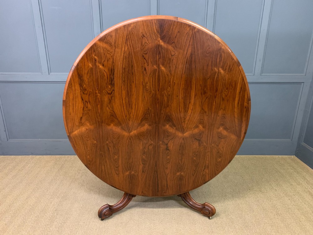 An oval Victorian rosewood loo table featuring a book-matched veneered top, crossbanded edge, and a carved central pedestal with four splayed legs on castors. View X
