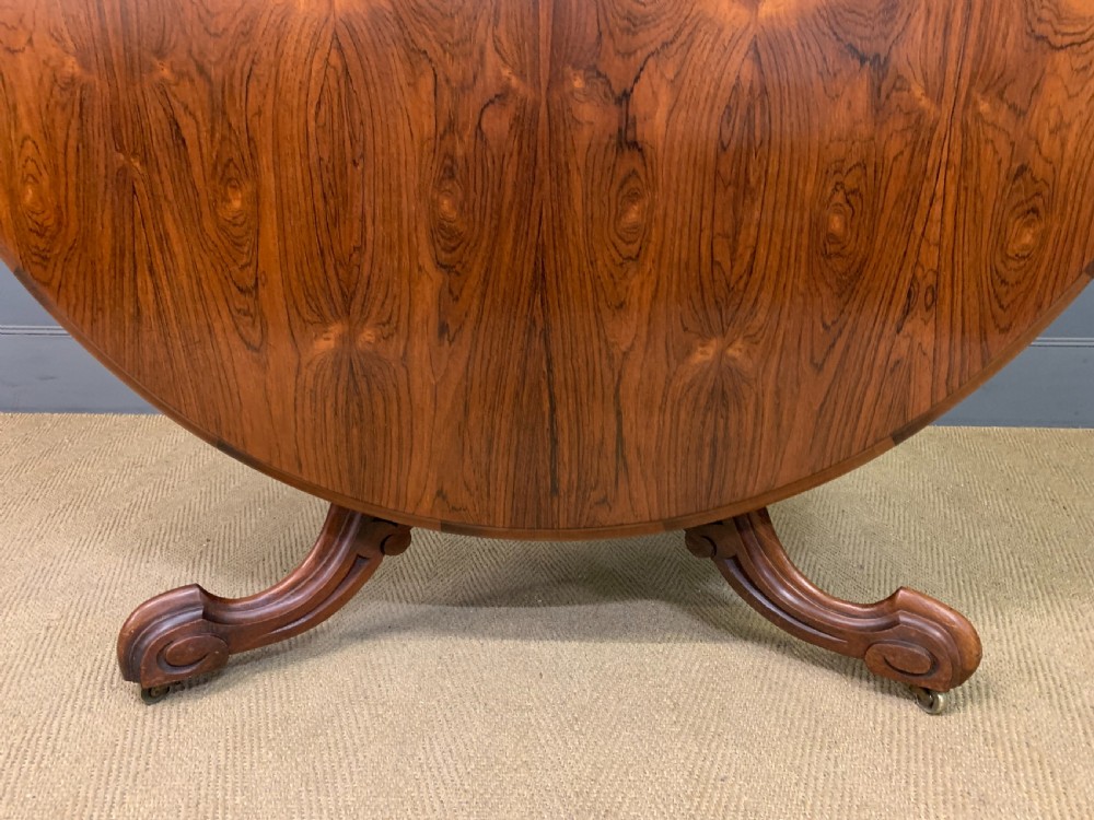 An oval Victorian rosewood loo table featuring a book-matched veneered top, crossbanded edge, and a carved central pedestal with four splayed legs on castors. View XII