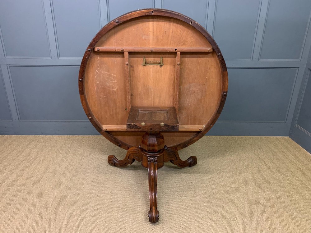An oval Victorian rosewood loo table featuring a book-matched veneered top, crossbanded edge, and a carved central pedestal with four splayed legs on castors. View XV
