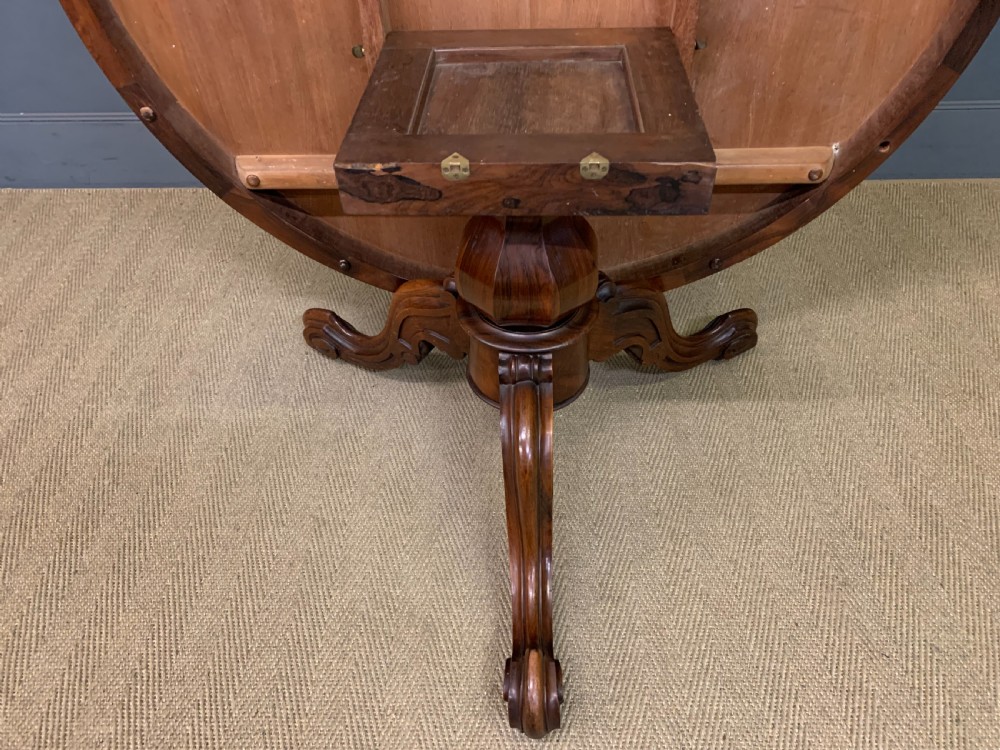 An oval Victorian rosewood loo table featuring a book-matched veneered top, crossbanded edge, and a carved central pedestal with four splayed legs on castors. View XVI