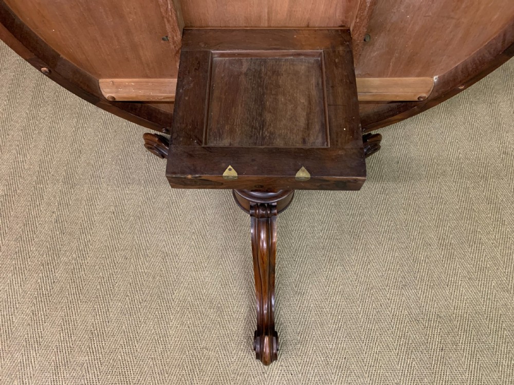 An oval Victorian rosewood loo table featuring a book-matched veneered top, crossbanded edge, and a carved central pedestal with four splayed legs on castors. View XVIII