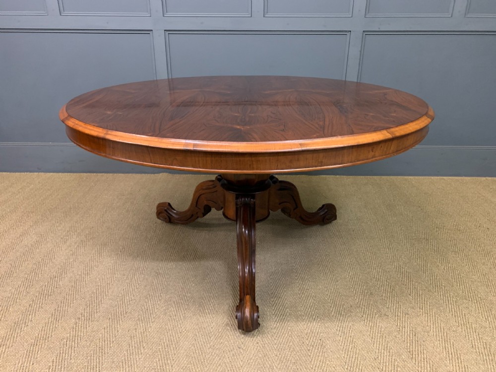 An oval Victorian rosewood loo table featuring a book-matched veneered top, crossbanded edge, and a carved central pedestal with four splayed legs on castors. View XIX