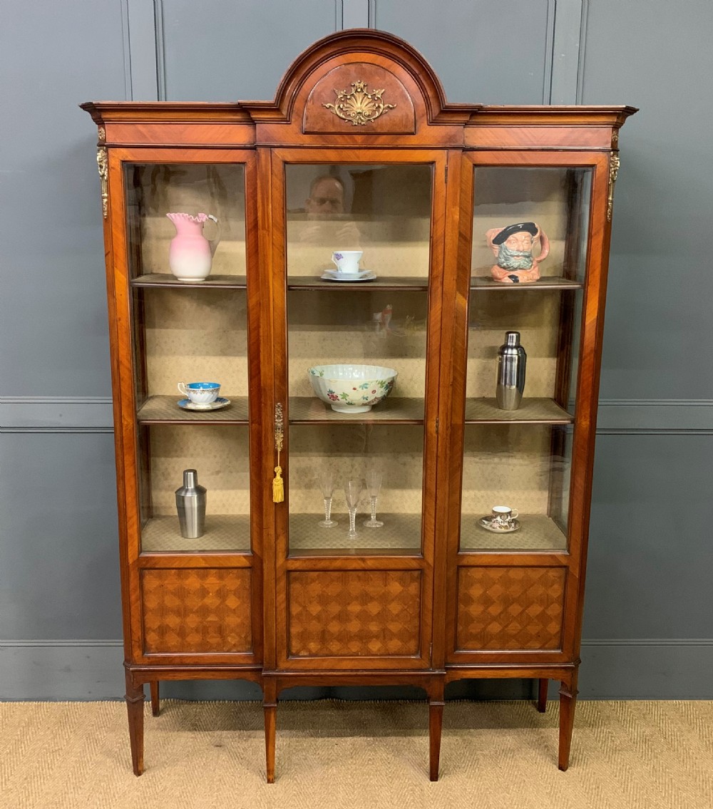 An Edwardian walnut and kingwood display cabinet featuring three glazed doors, parquetry panel lower sections, and a central arched pediment with a gilt mount.
