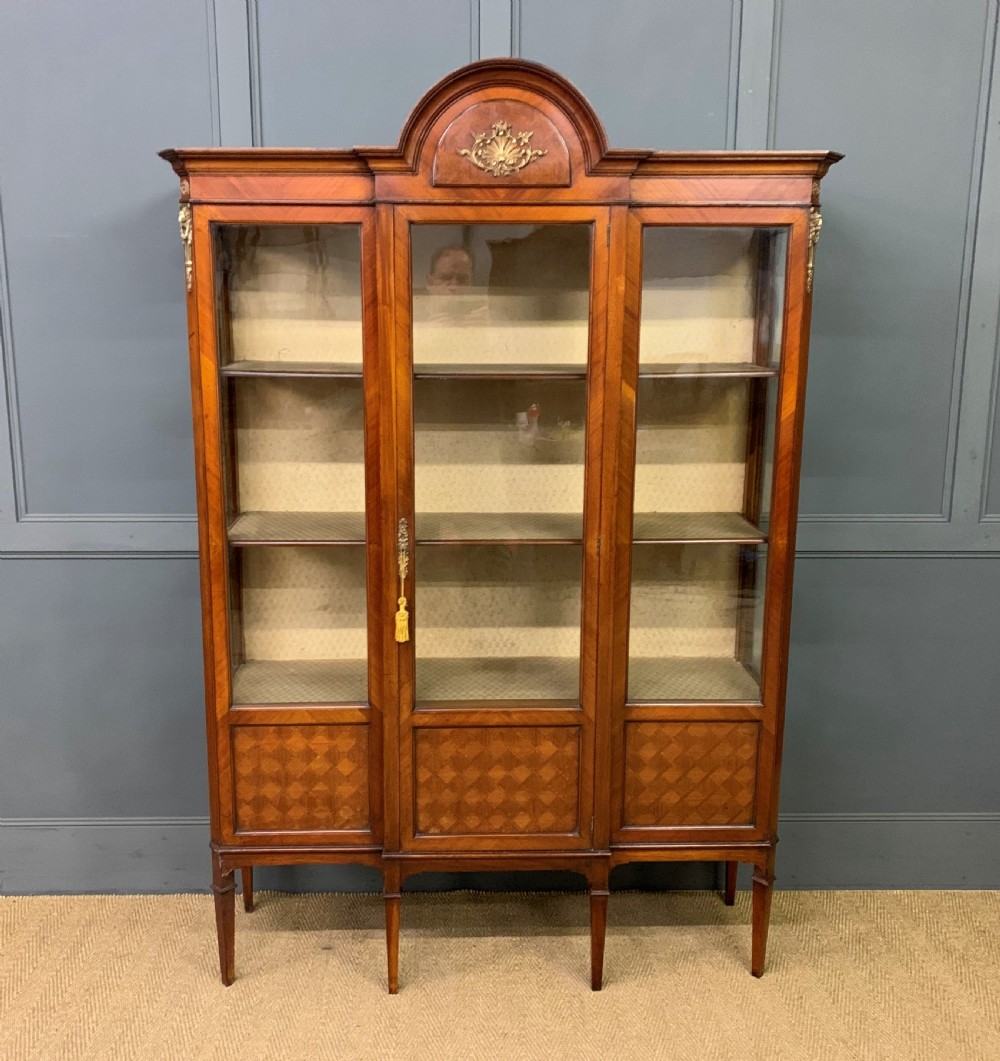 An Edwardian walnut and kingwood display cabinet featuring three glazed doors, parquetry panel lower sections, and a central arched pediment with a gilt mount. View II