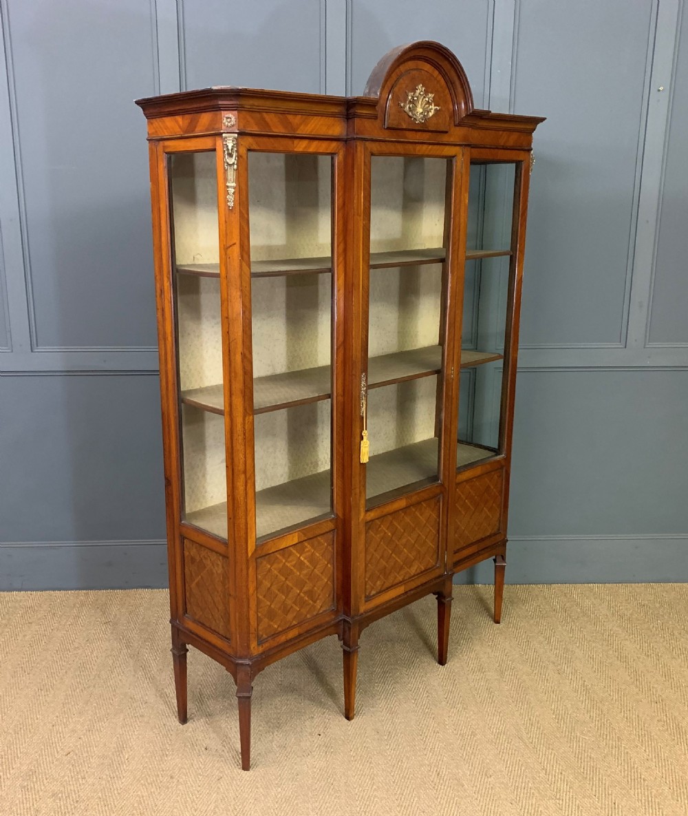 An Edwardian walnut and kingwood display cabinet featuring three glazed doors, parquetry panel lower sections, and a central arched pediment with a gilt mount. View VIII