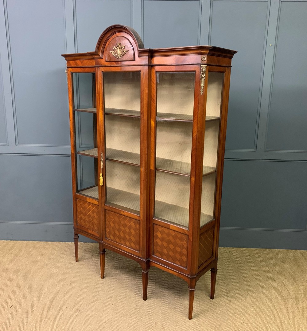 An Edwardian walnut and kingwood display cabinet featuring three glazed doors, parquetry panel lower sections, and a central arched pediment with a gilt mount. View XIV