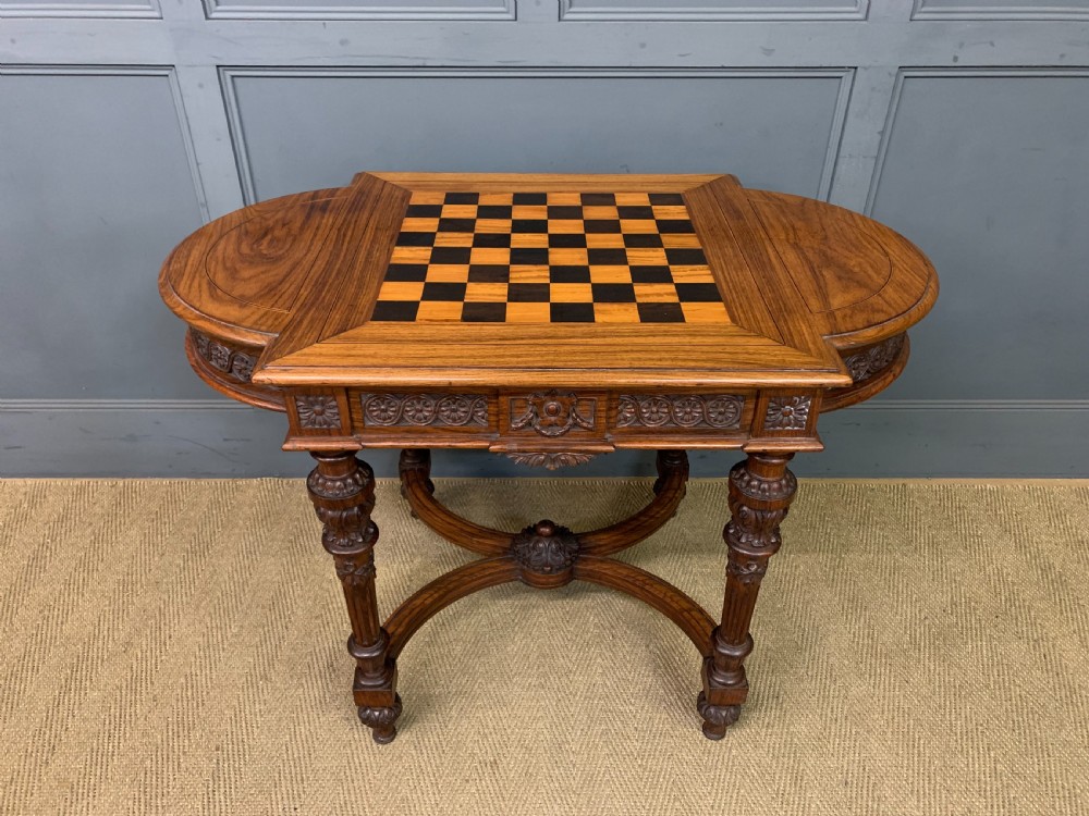 An Anglo-Indian rosewood games table featuring an inlaid square chessboard top, drop leaves, a carved frieze, and turned legs connected by a curved stretcher.