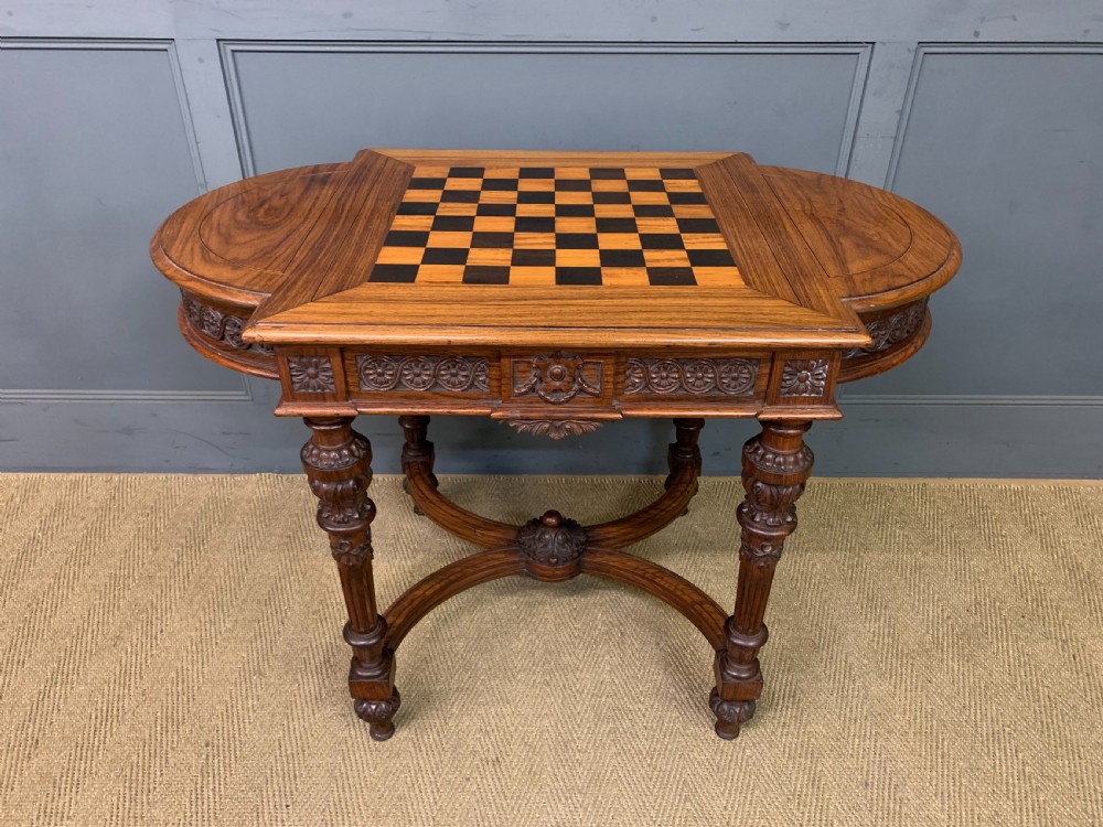 An Anglo-Indian rosewood games table featuring an inlaid square chessboard top, drop leaves, a carved frieze, and turned legs connected by a curved stretcher. View II