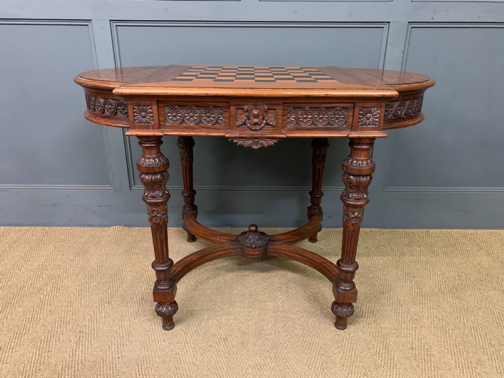 An Anglo-Indian rosewood games table featuring an inlaid square chessboard top, drop leaves, a carved frieze, and turned legs connected by a curved stretcher. View III